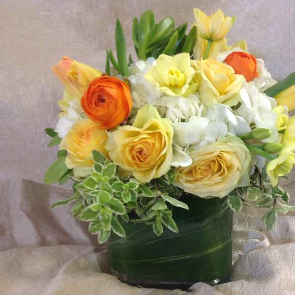 Compact arrangement of yellow roses, white hydrangeas, and orange blooms in a leaf-wrapped glass vase.