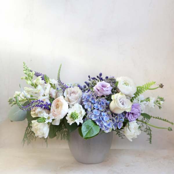 Low floral arrangement in a gray vase with white, lavender, and blue blooms