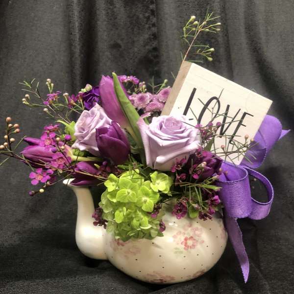 Purple and lavender floral arrangement in a white ceramic vase with a ribbon and "LOVE" sign