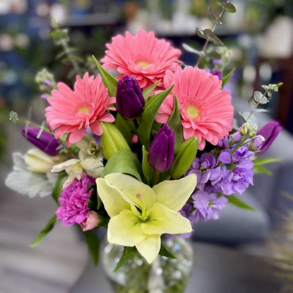 Mixed bouquet of pink gerbera daisies, purple tulips, and a pale lily in a glass vase