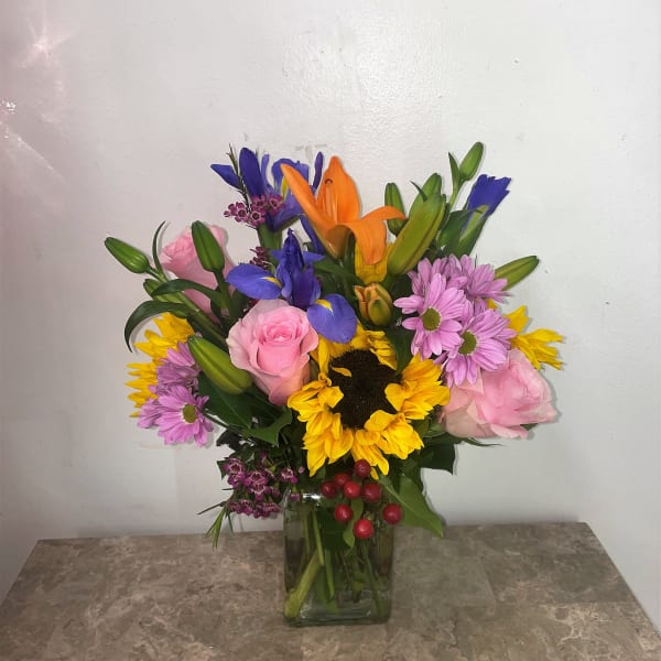 Mixed bouquet with sunflowers, roses, lilies, and purple daisies in a glass vase