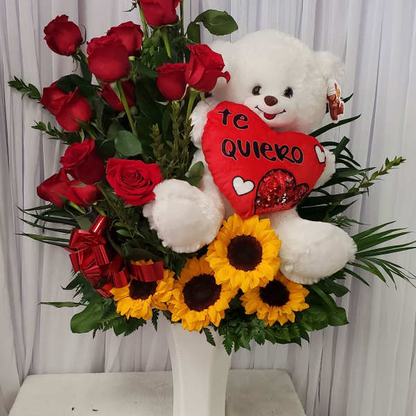 Red roses and sunflowers arranged with a teddy bear and heart pillow in a white vase.