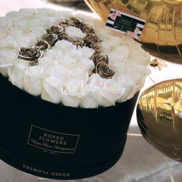 White roses arranged in a black round box with gold balloons nearby.