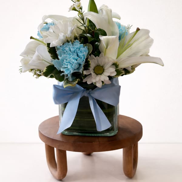White lilies and daisies with blue carnations in a glass vase with a blue ribbon