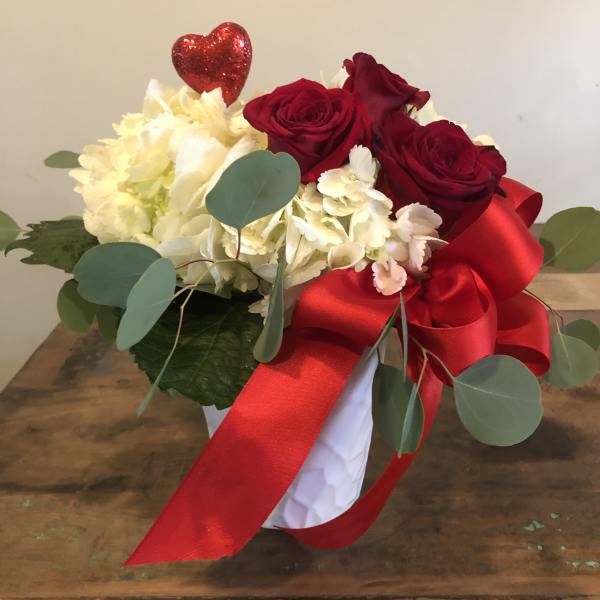 Red roses and white flowers in a white vase with a red ribbon and heart pick