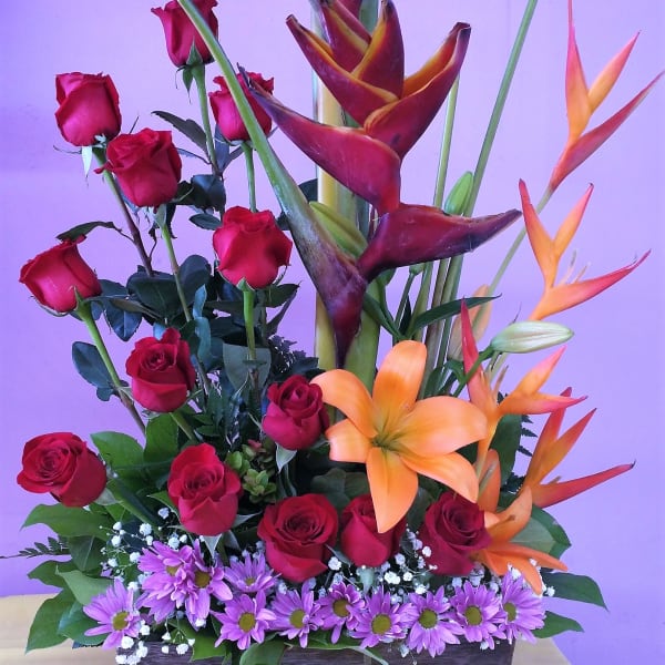 Tall mixed flower arrangement with red roses, orange heliconia, and purple daisies in a rectangular container