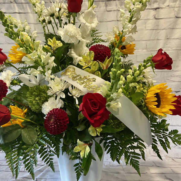 Red roses, sunflowers, and white flowers in a white vase with a ribbon