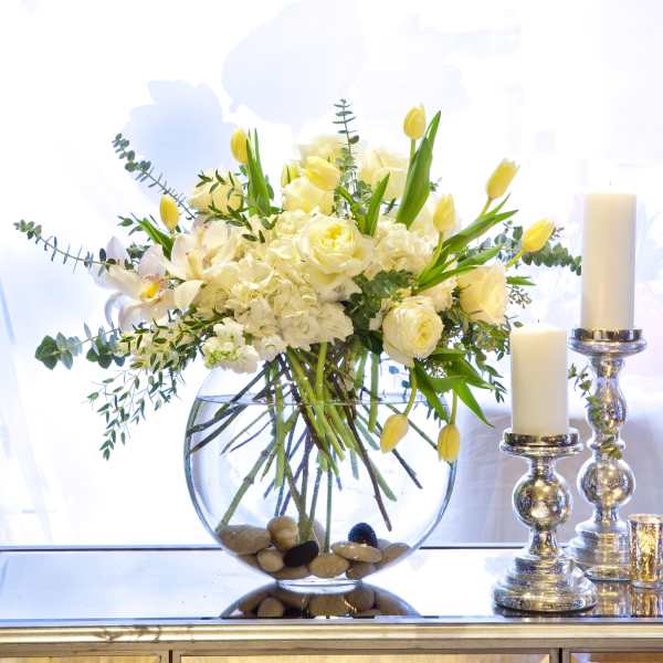 White and yellow floral arrangement in a round glass vase beside candles
