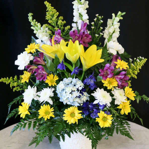 Mixed floral arrangement with yellow lilies, white blooms, and purple flowers in a white vase