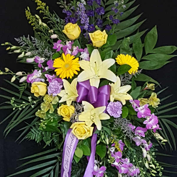 Standing floral spray with yellow roses, lilies, and purple flowers on a black background