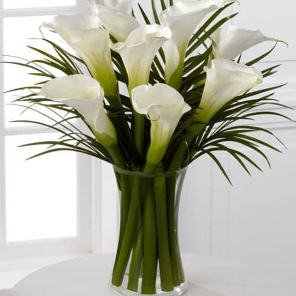 Tall arrangement of white calla lilies with long blades of foliage in a clear glass vase
