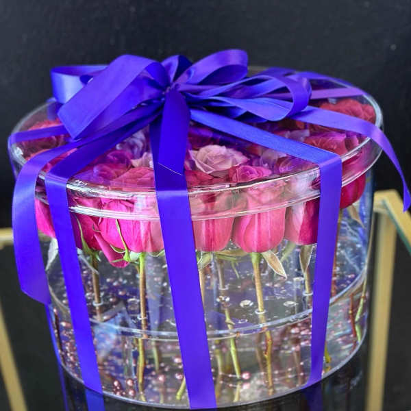 Pink roses in a clear round box tied with a purple ribbon