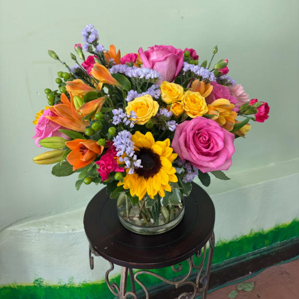 Mixed bouquet of roses, sunflowers, lilies, and small purple filler flowers in a glass vase