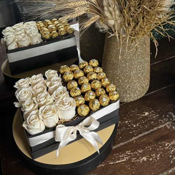 White roses and gold chocolates in black gift boxes beside a glittery vase of dried stems