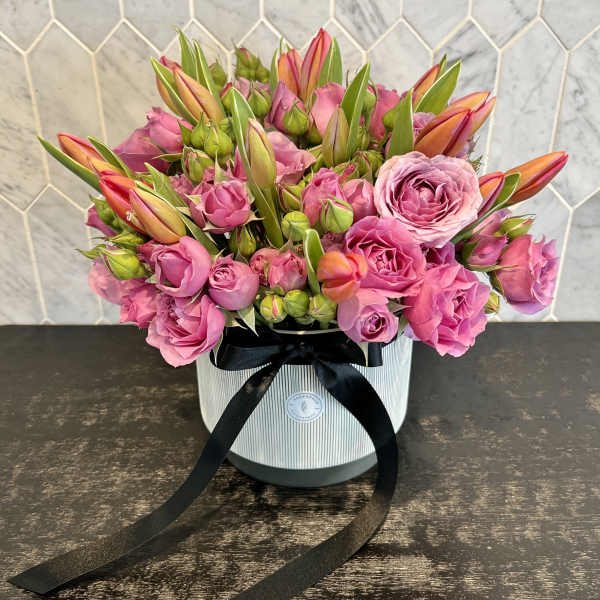 Pink roses and orange tulips arranged in a striped hat box with a black ribbon.