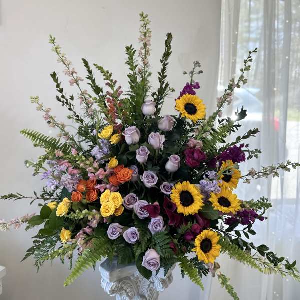Large mixed floral arrangement with sunflowers, roses, and purple blooms in a pedestal vase