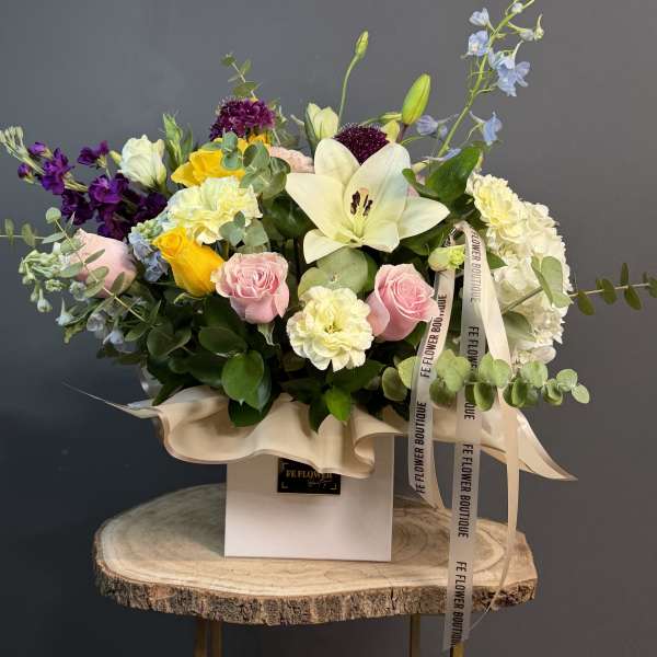 Colorful mixed flower arrangement with lilies, roses, and carnations in a white box on a wood stand.