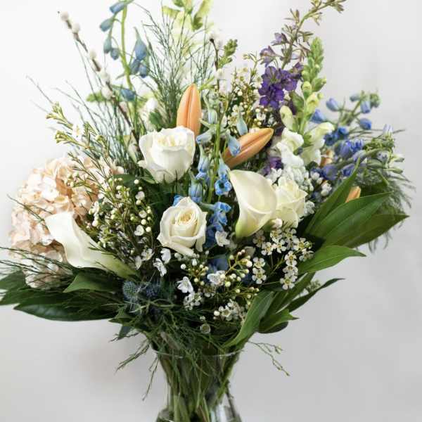 Tall mixed bouquet of white and pastel flowers with blue accents in a clear glass vase