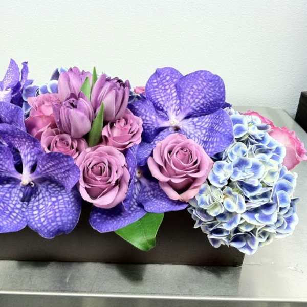 Purple roses and orchids arranged in a rectangular box with blue hydrangeas