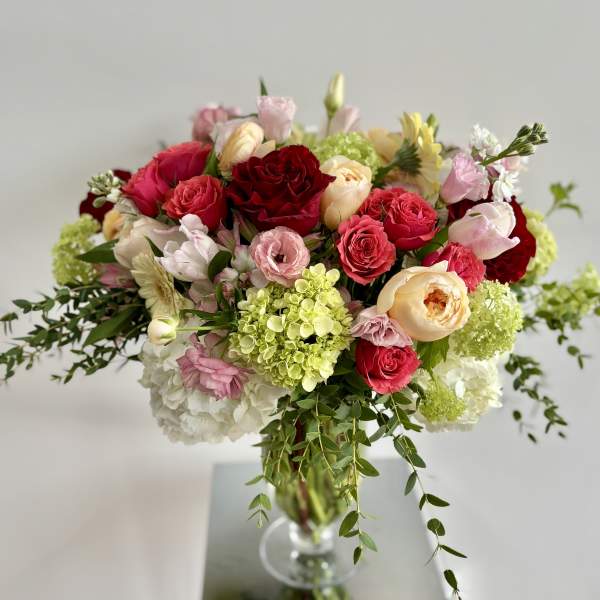 Mixed arrangement of red, pink, and peach roses with green hydrangeas in a clear glass vase