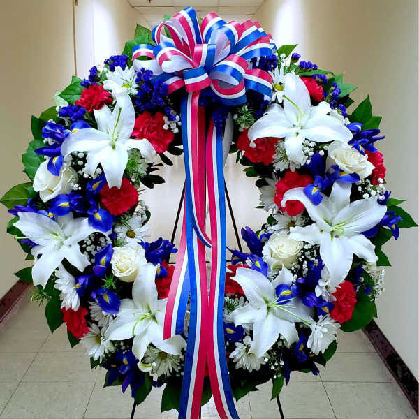 Large floral wreath with white lilies, red carnations, blue flowers, and a tricolor ribbon bow