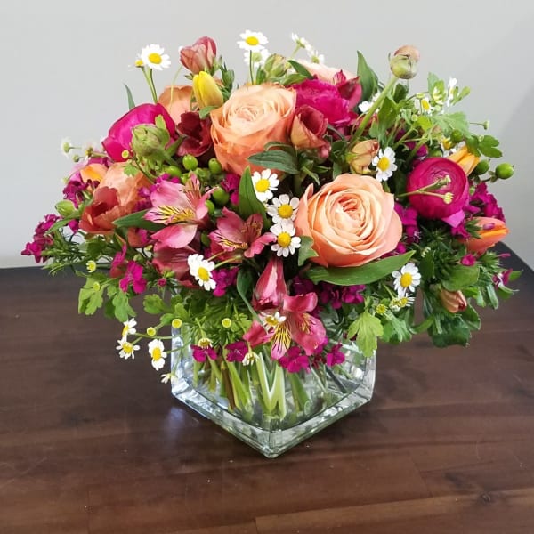 Mixed bouquet of peach roses, pink flowers, and small white daisies in a clear glass cube vase.