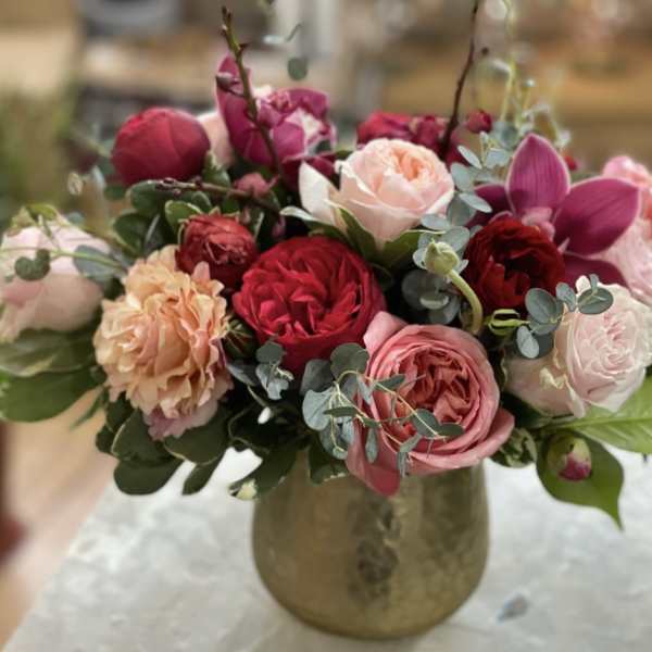 Low gold vase with pink, red, and peach roses and orchids arranged in a lush rounded bouquet.