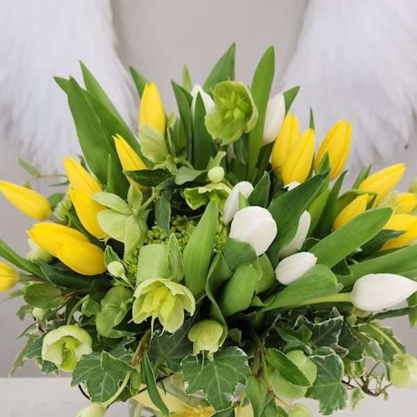Yellow and white tulip arrangement with green blooms in a round vase with ivy and a yellow ribbon bow.