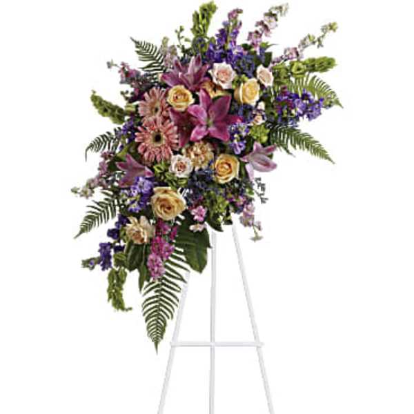 Standing sympathy spray with pink lilies, roses, and mixed purple and yellow flowers on a white easel