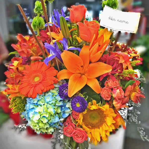 Bright mixed bouquet with orange lilies, roses, sunflowers, and blue hydrangea in a glass vase