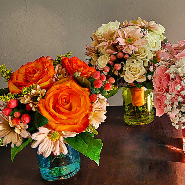 Three small mixed flower arrangements with roses and daisies in colored glass jars on a table.