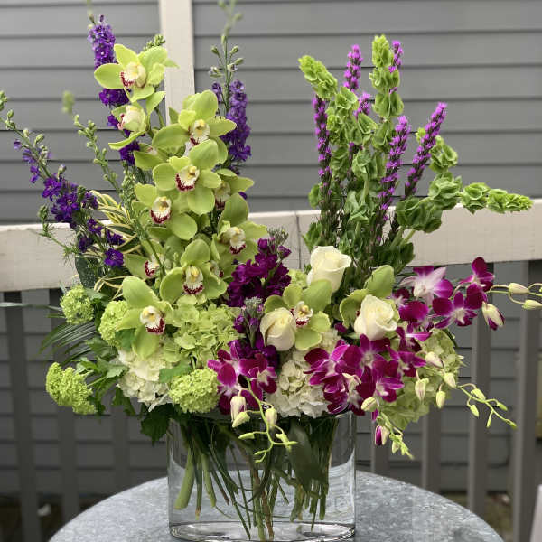 Tall floral arrangement with green orchids, purple blooms, and white roses in a glass vase
