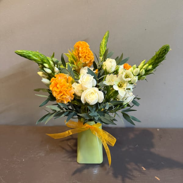 White roses and orange blooms in a green vase with a yellow ribbon