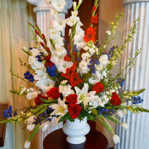 Tall red, white, and blue floral arrangement with roses, lilies, and delphinium in a white urn vase