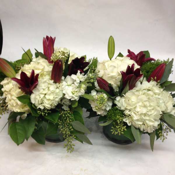 Two white floral arrangements with burgundy lilies in dark containers