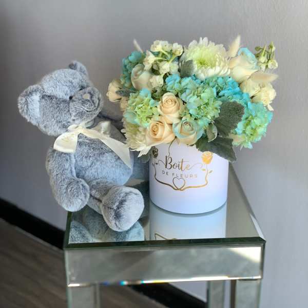 Blue teddy bear beside a pastel flower arrangement in a white box