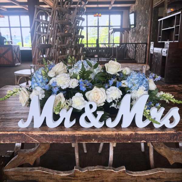 Low wedding centerpiece of white and blue flowers with a white Mr & Mrs sign on a rustic wooden table