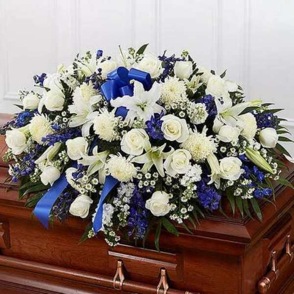 White and blue funeral casket spray with roses, lilies, and ribbon on a wooden casket