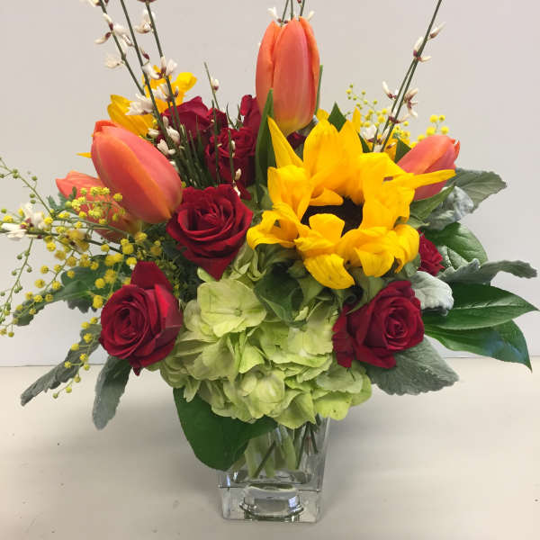 Mixed bouquet with red roses, yellow sunflowers, and orange tulips in a clear vase