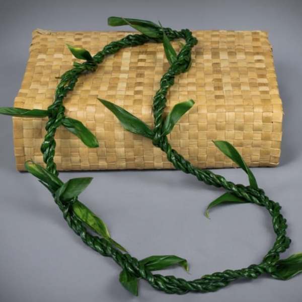 Braided green leaf lei draped over a tan woven mat against a gray background
