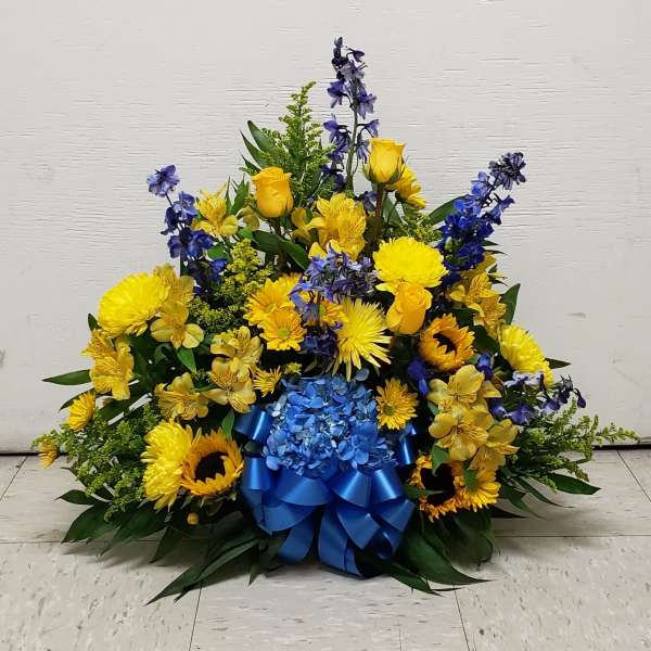 Fan-shaped yellow and blue arrangement with sunflowers, roses, delphinium, and a blue ribbon bow.