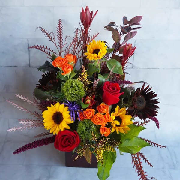 Mixed bouquet with sunflowers, roses, and orange spray roses in a square vase