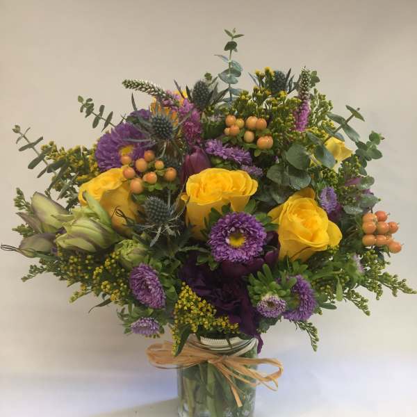 Bright yellow roses and purple asters arranged with berries in a mason jar tied with raffia