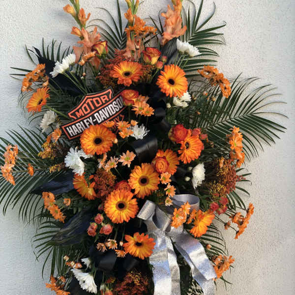 Tall standing spray with orange gerbera daisies, white flowers, palm fronds, and a Harley-Davidson logo sign.