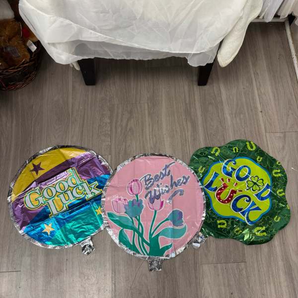 Three round foil balloons on a floor, reading Good Luck, Best Wishes, and Good Luck.