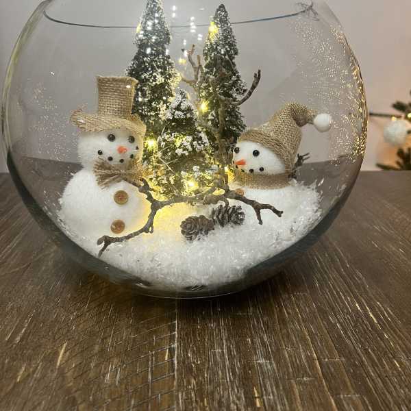 Two snowman figurines in a glass bowl with miniature trees and lights
