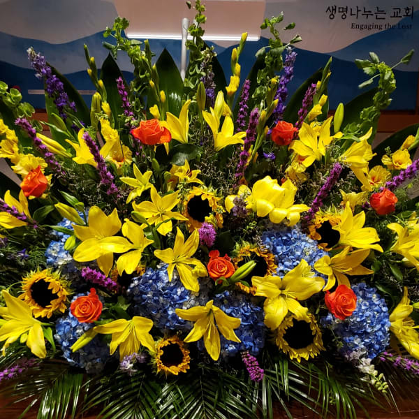 Large floral arrangement with yellow lilies, blue hydrangeas, and orange roses