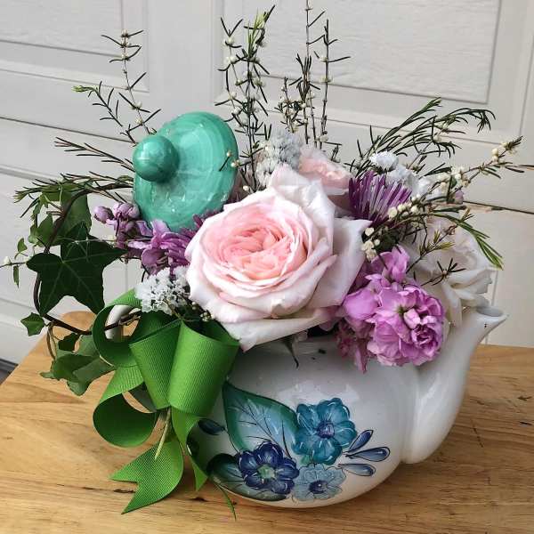 Pastel pink and purple flower arrangement in a painted ceramic teapot with green bow and tilted lid