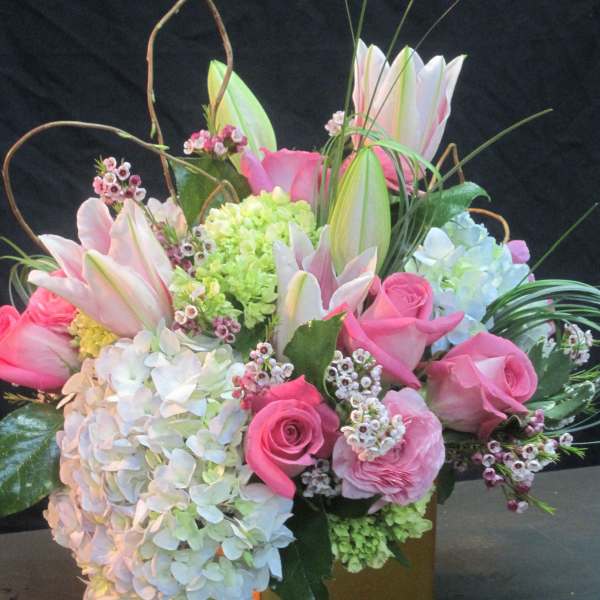Compact arrangement of pink roses, white hydrangeas, and lilies in a gold cube vase