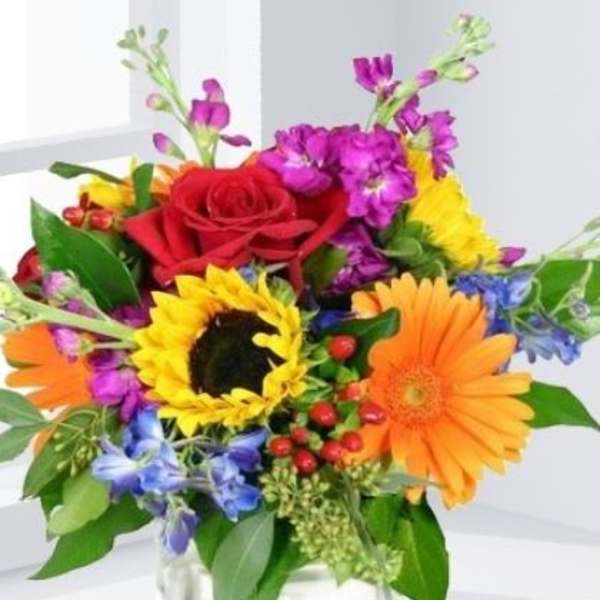 Colorful mixed bouquet in a clear square vase with a red rose, sunflower, and gerbera daisies
